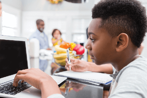  A photograph of a middle school student working at a laptop at his desk while taking notes.