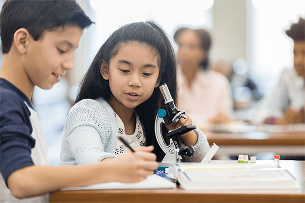 Two students sit at a table in a science classroom in front of a microscope discussing their findings and one student records the findings in his notebook.