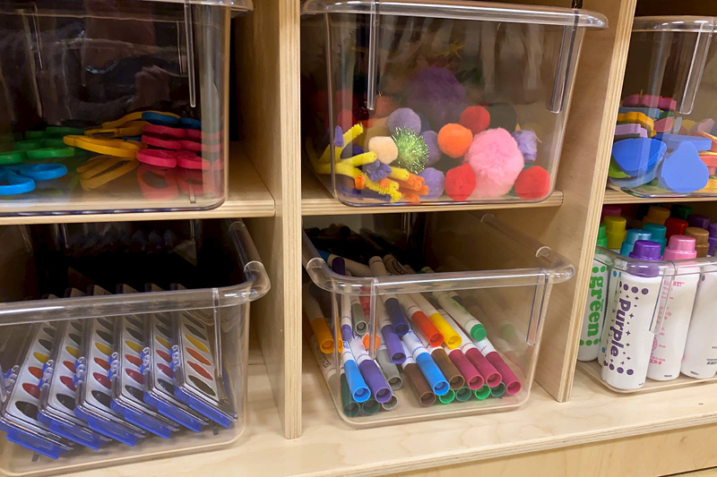 Clear bins organized on a classroom shelf with various supplies in each one.