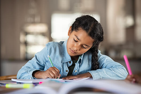 An elementary-aged student concentrates on a writing assignment at her desk in a classroom.