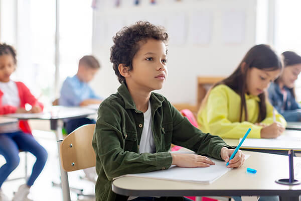 A young student sitting at a desk in a classroom listening to the teacher during a writing lesson.