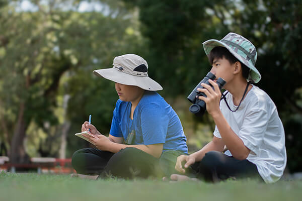 Two students sit side by side in an outdoor area like a park, one observing nature with binoculars and the other taking notes, as part of an inquiry-based science lesson.