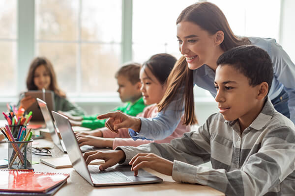 A smiling teacher assists a boy at his desk working on a laptop in a classroom while other students engage in other classroom activities.