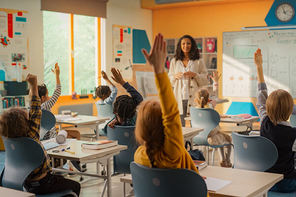 A classroom of young students are eagerly raising their hands while the teacher stands in front of them smiling during a literacy lesson.