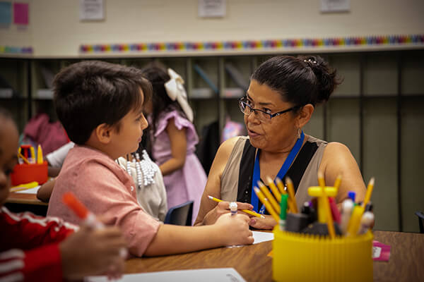 A Kansas City, Kansas Public Schools teacher works with a young student at a desk in a classroom.
