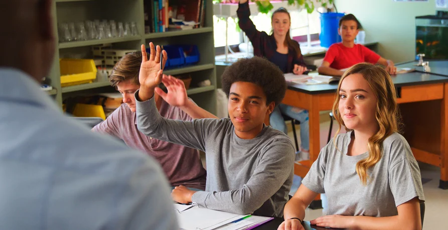Photograph of high school students sitting at tables in a classroom. Several students are raising their hands. Image used as an example of the “Share New Learning” reading comprehension practice activity.