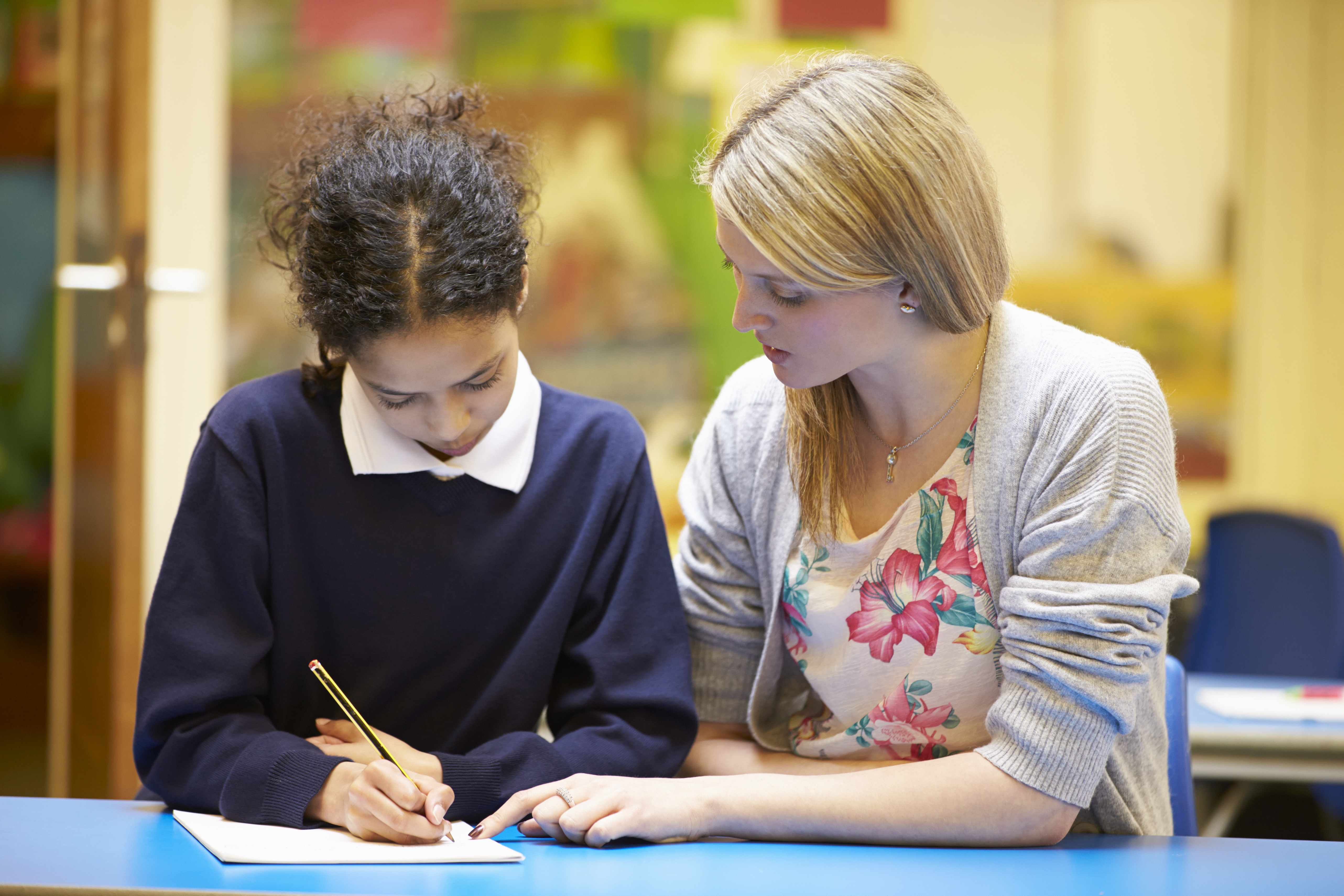 A teacher sitting alongside a middle school student at a table with a book in front of them helping her with her reading. 