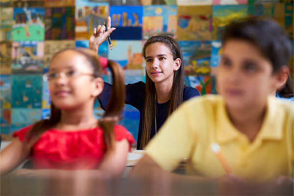 A young student is raising her hand in a social studies classroom to answer a big question asked by a teacher to prompt an inquiry-based learning activity.