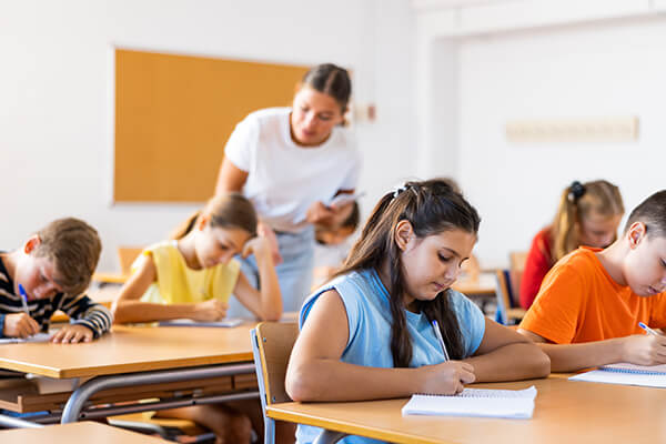 A classroom of middle grade students are engaged in a writing assignment at their desk while a teacher checks in with individuals during a lesson.
