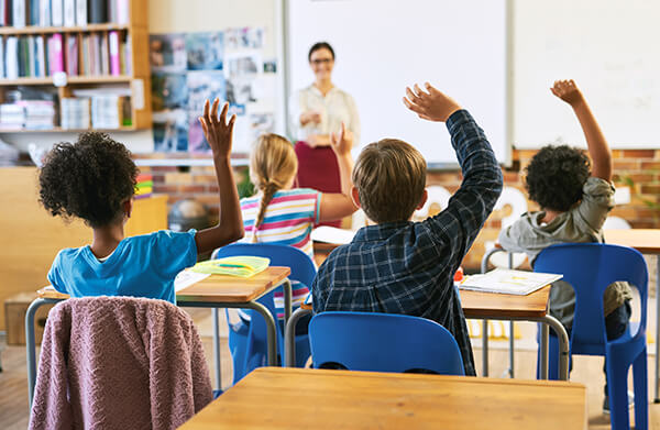 Students enthusiastically raising their hands in a classroom with a teacher who is happily teaching at the front of the class.