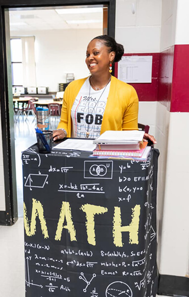 A Clayton County Public Schools math teacher stands, smiling outside her classroom at a podium with a banner across the front that reads, “math.”