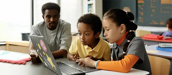 elementary-students-doing-assignment-on-laptop-with-teacher-705x310.webp