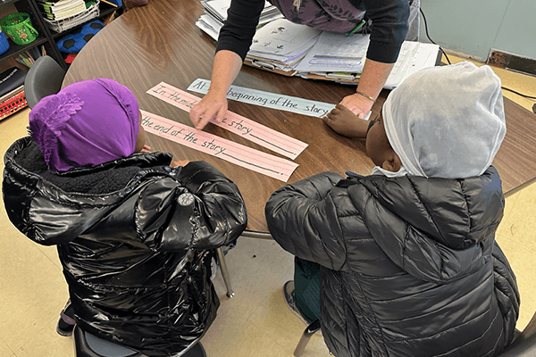 Two Lewiston Public School students sitting side-by-side at a table looking at sentence strips while a teacher explains the lesson in front of them.