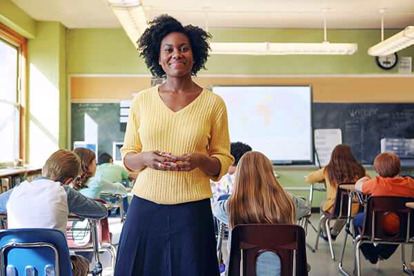 A teacher stands in a classroom with students feeling proud of the work she has done with her part in successfully implementing a new curriculum.