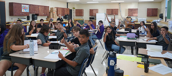 A mix of middle school students confidently giving a thumbs up in a classroom setting.