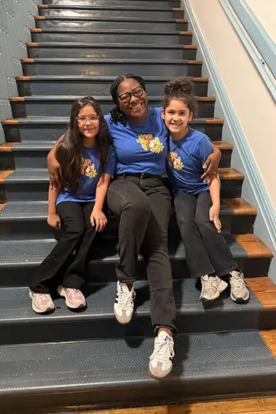 A teacher from Susie King Taylor Community School sitting in a stairwell smiling next to two happy elementary-aged students.