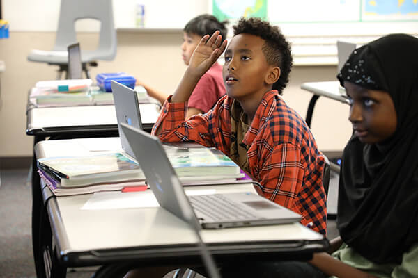 Seattle Public Schools student in a classroom raising his hand and looking engaged in what the teacher is saying at the front of the room.