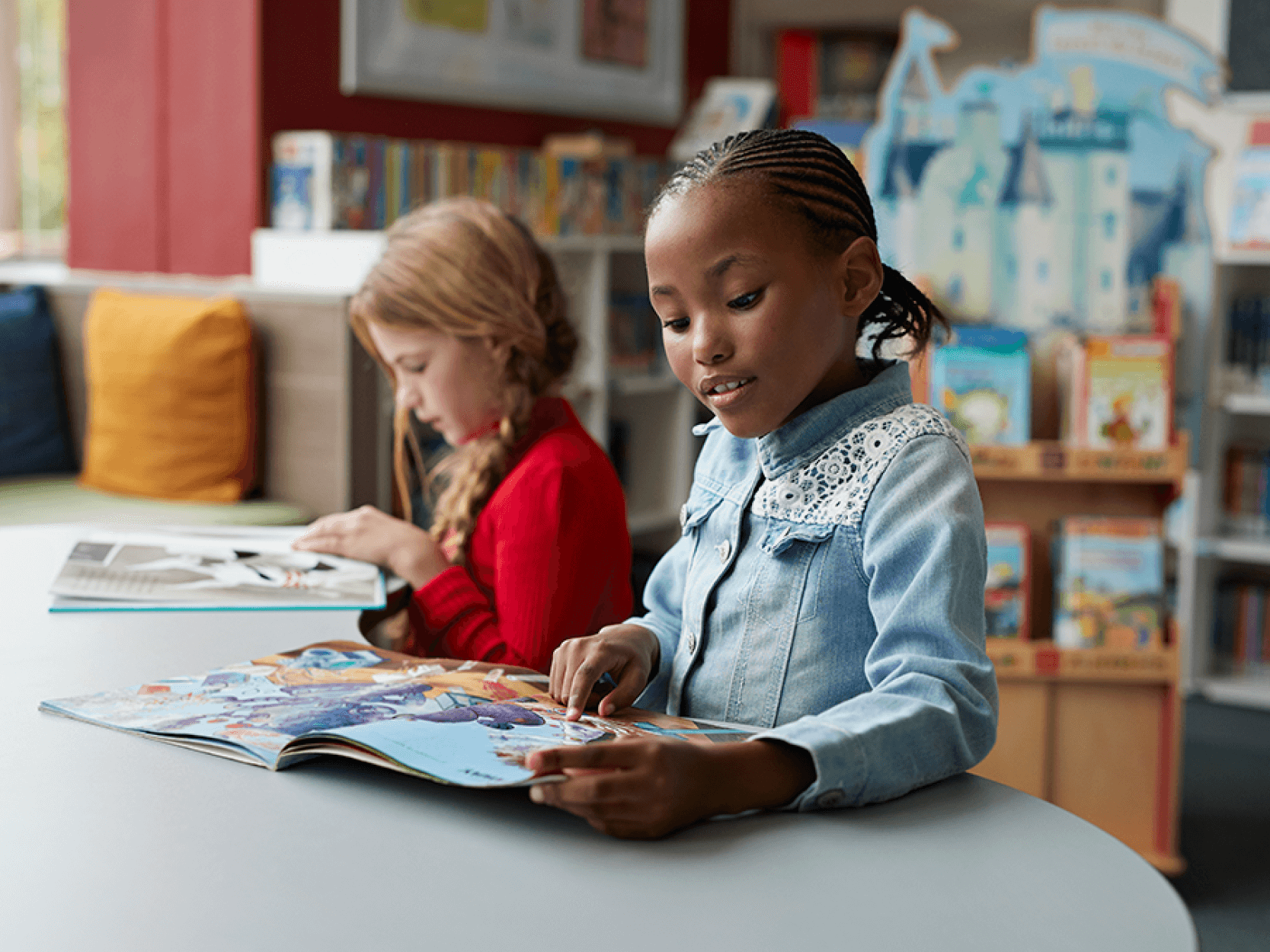 Two young students sit next to each other at a table reading different kinds of books.