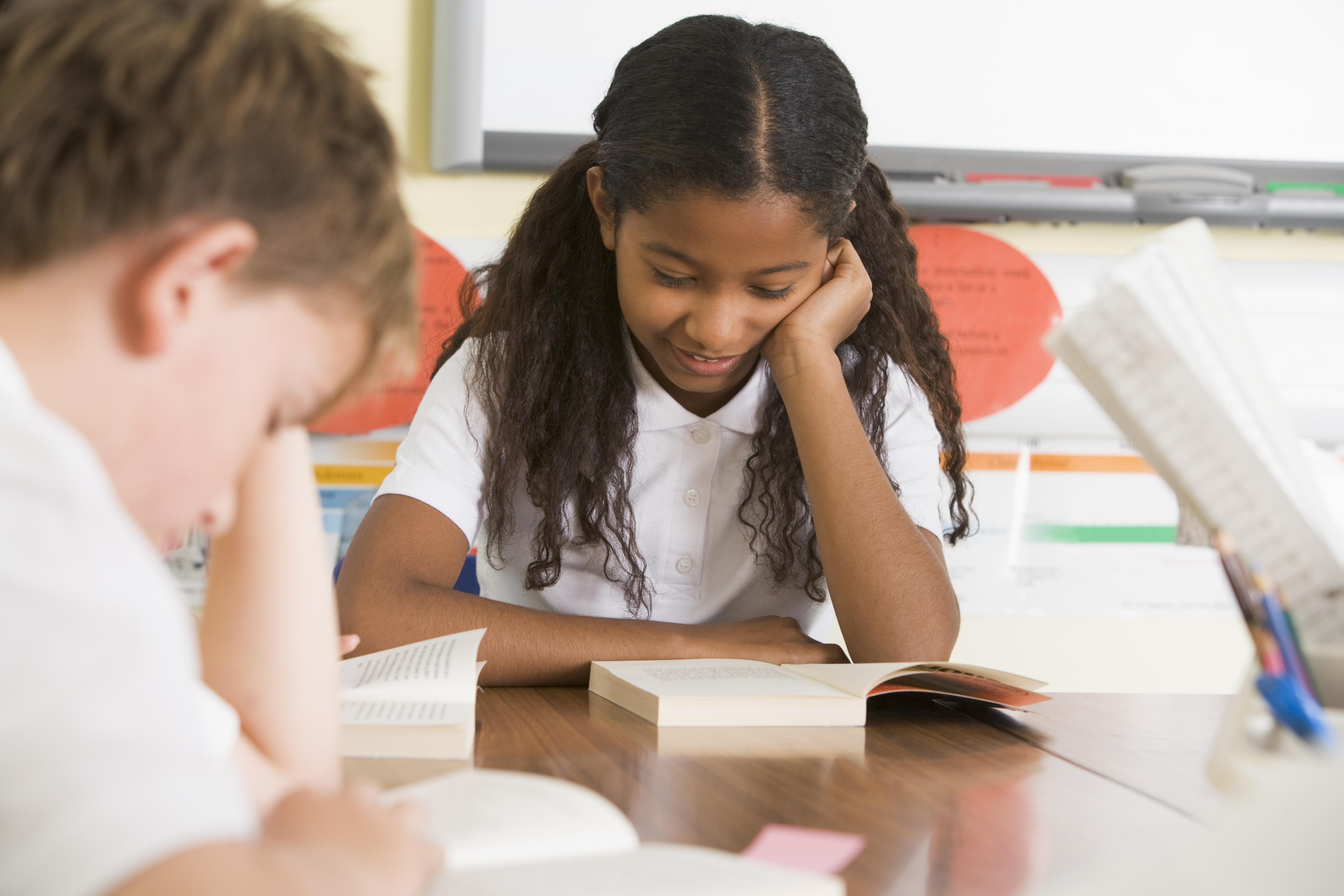 A middle school student sitting at a desk smiling slightly and engaged with a book she is reading in front of her.