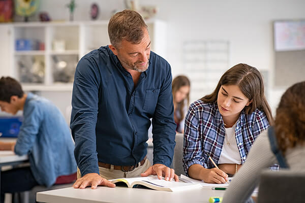 A teacher standing over a middle school student sitting at her desk while the teacher models fluent reading to her. 