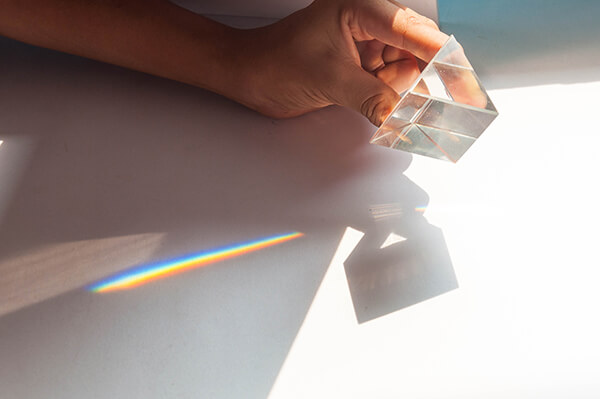 A hand holds a crystal prism in the light so that the prism casts a rainbow on a flat surface.