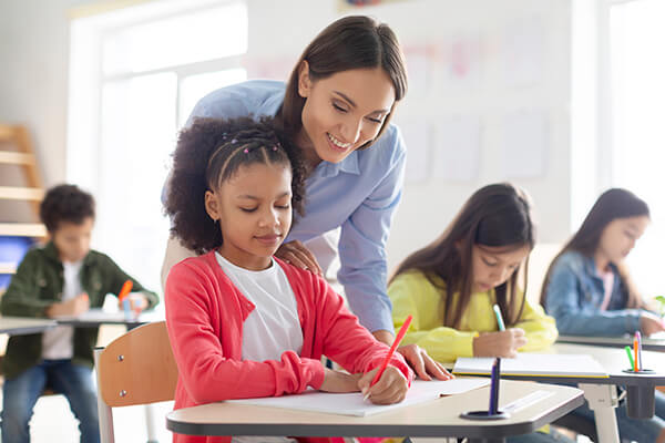 A cheerful teacher helps an elementary-aged student with her writing assignment. 