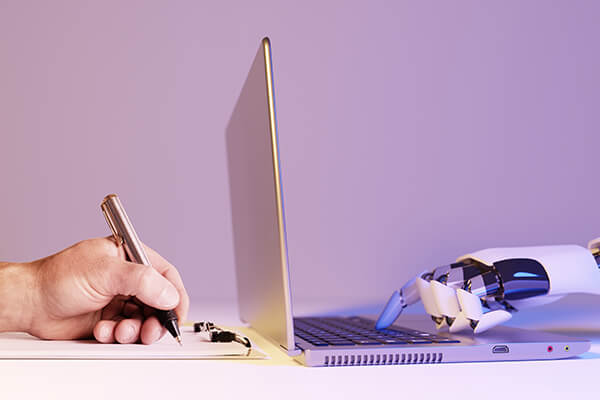 Two laptops lay back to back on a desktop with a human hand typing on the left, while a robot hand types on the left.