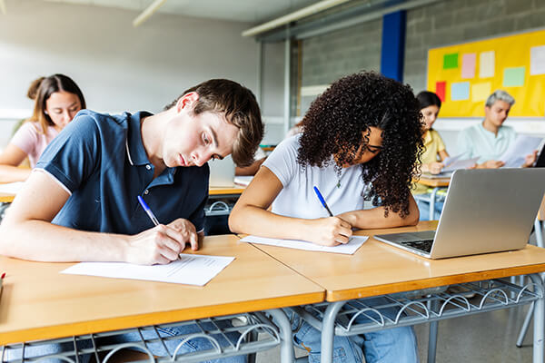 A group of students in a classroom is focused on writing their assignments at desks, with a young man and a young woman sitting in the foreground, while others work in the background.