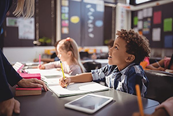 A photograph of a young student at his desk in a classroom looking up and listening to the teacher talk to him about a writing assignment.